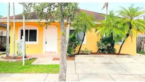 A single-story house with a yellow exterior, a front porch, and a small front yard with a palm tree. - Homestead Green retreat (Homestead)