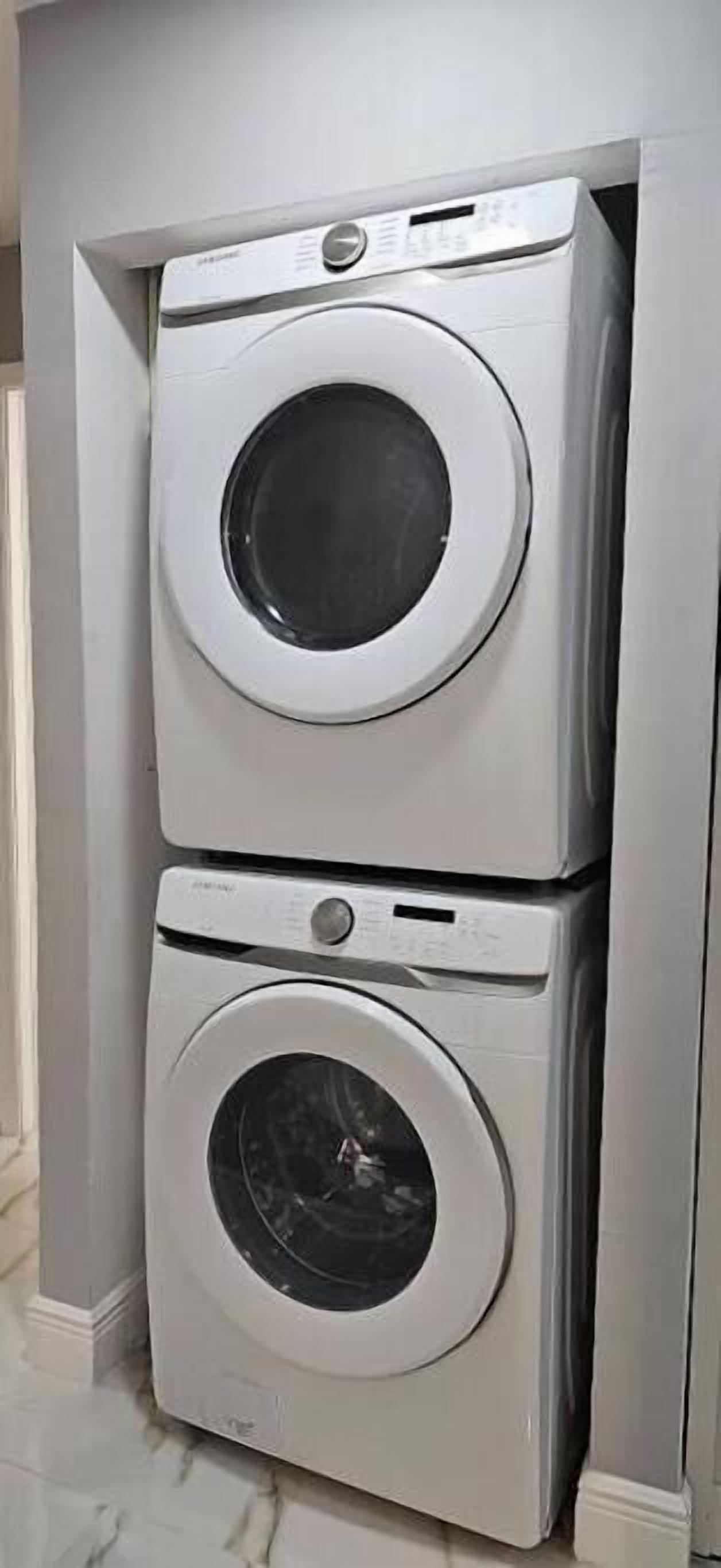 A stacked washer and dryer unit in a corner of a room with a marble floor.