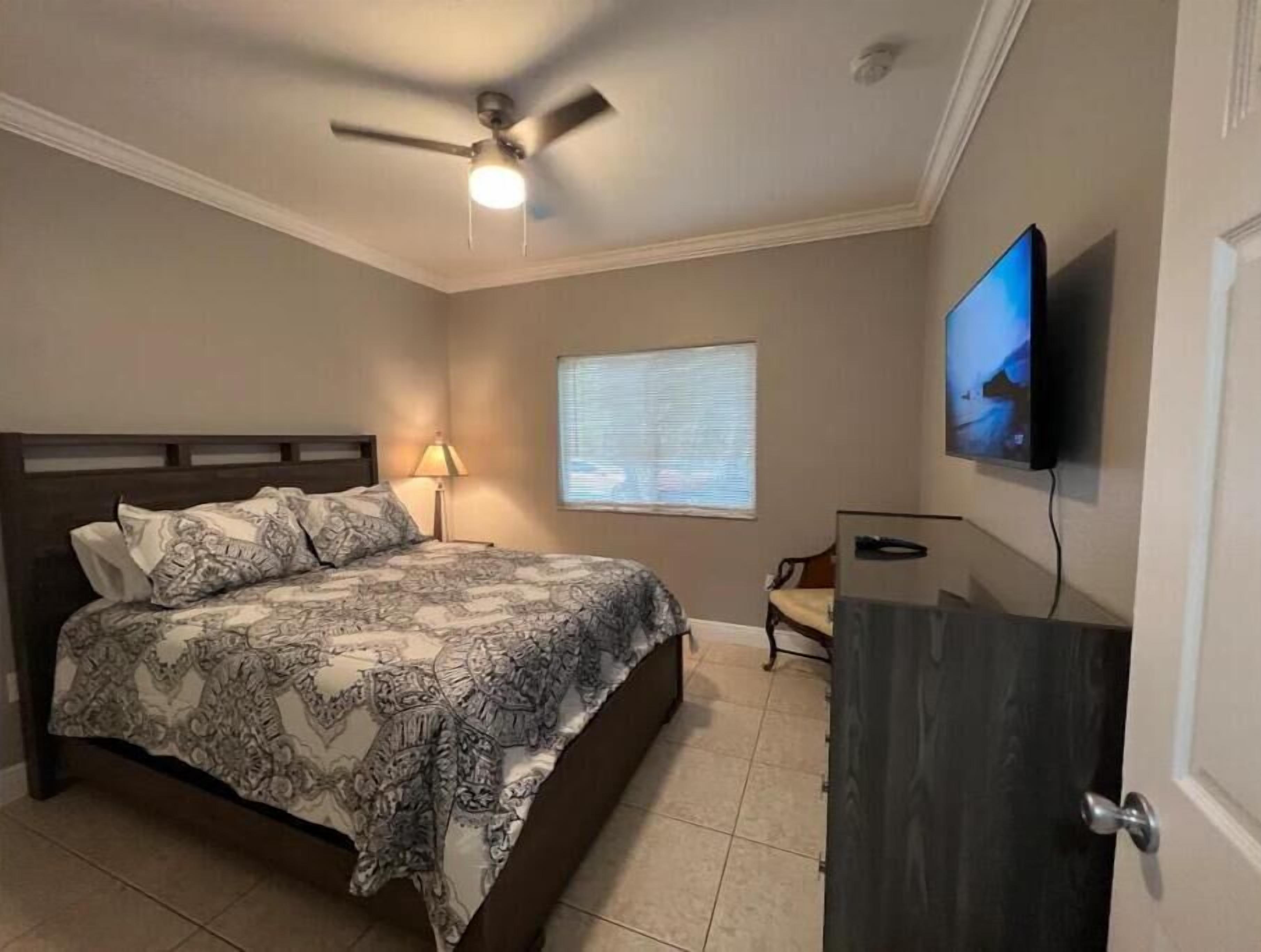 Un dormitorio con cama, ventilador de techo, televisor montado en la pared y ventana.