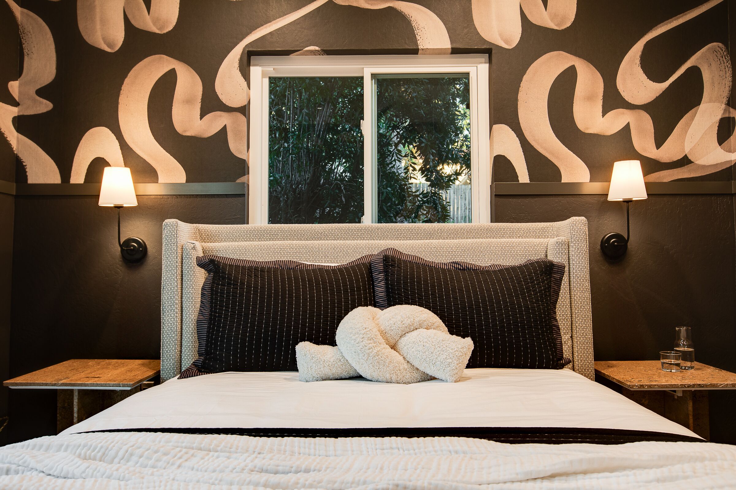 Een hotelkamer met een groot bed, twee bedlampjes, een raam met uitzicht op bomen en een kamer met een patroon op de muur.