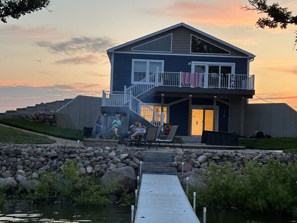 A two-story house with a balcony, a dock leading to a body of water, and a stone wall. - Country lake house getaway accommodating large family-sized fun! (Emmetsburg)
