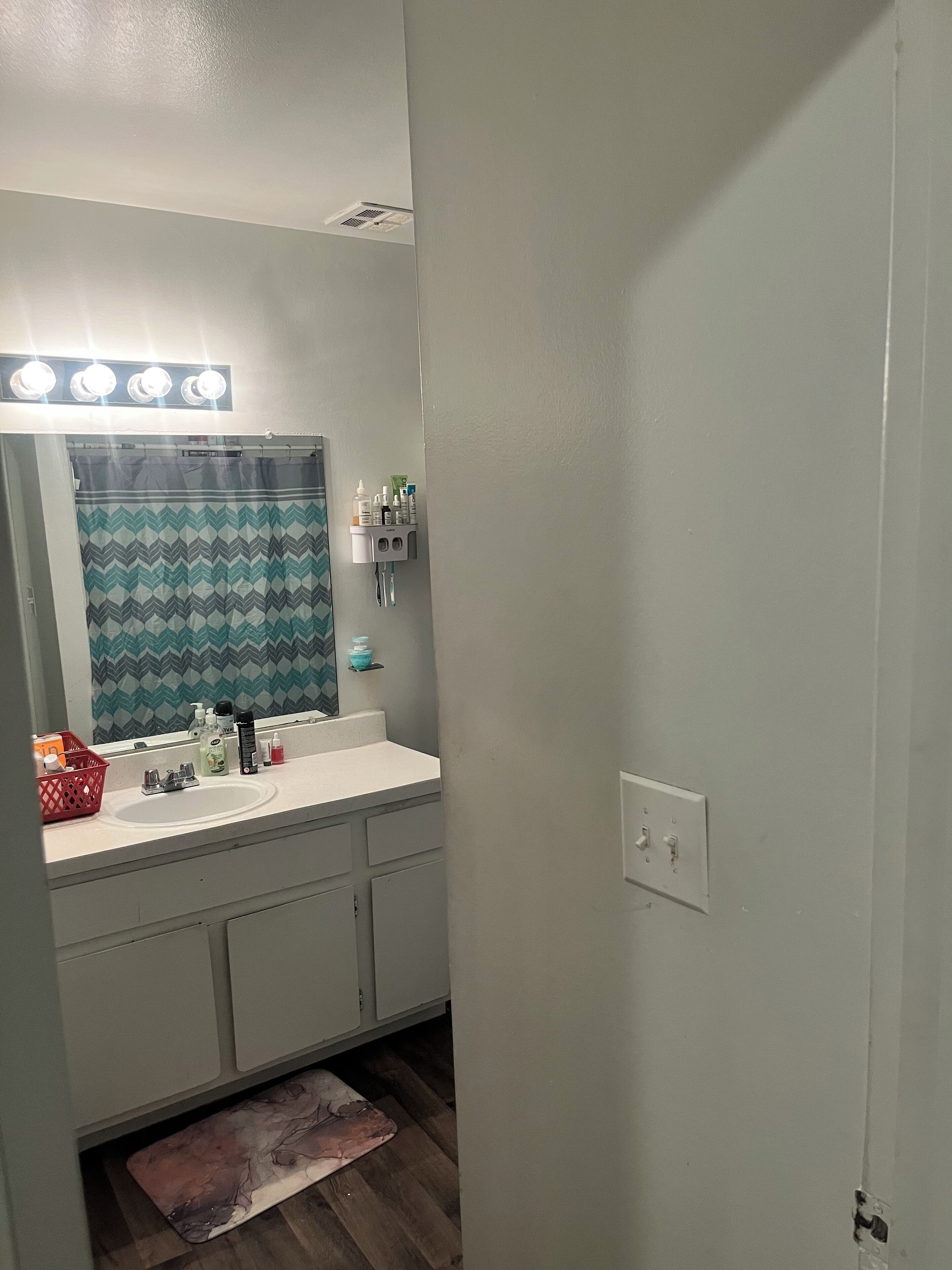 Un bagno con un lavandino bianco, uno specchio e una tenda da doccia. Sul lavandino ci sono articoli da toilette e un cesto rosso sul pavimento.