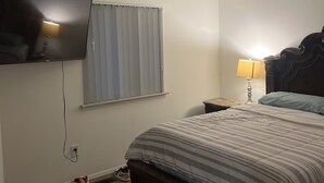 A bedroom with a bed, a TV mounted on the wall, a window with blinds, a bedside lamp, and a nightstand. - Beautiful get away right outside Detroit, peace of mind. Lots of attractions (Taylor)