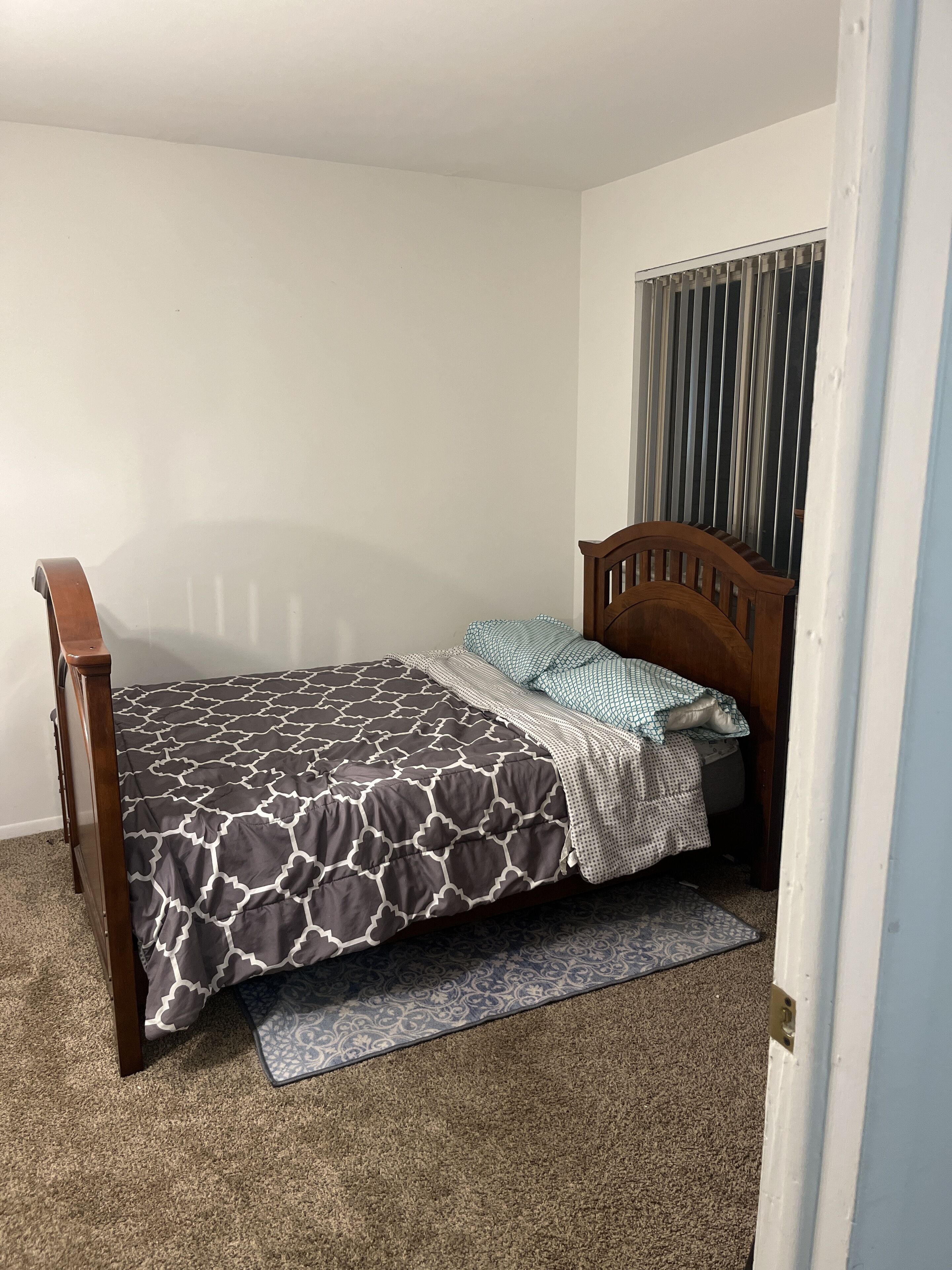 A bedroom with a wooden bed frame, a patterned bedspread, a blue and white pillow, and a window with vertical blinds.
