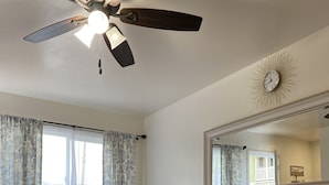 A living room with a ceiling fan, a large window with patterned curtains, a beige sofa with multiple cushions, and a wall-mounted mirror.