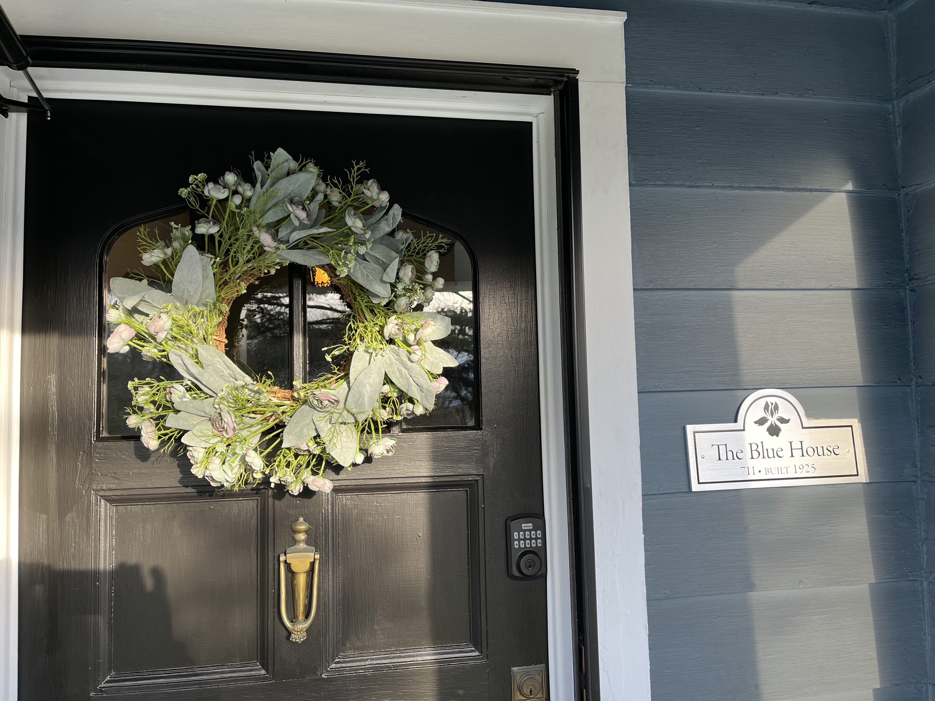 A black door with a wreath, a brass knocker, and a keypad lock. A sign on the wall reads "The Blue House 71, Bull 1925".