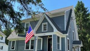 A two-story house with a gabled roof, a front porch, and a flagpole with an American flag. - Explore the Finger Lakes Region from this cozy 3-bedroom home near Geneva, NY. (Waterloo)