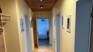 A hallway with wooden flooring, beige walls, and framed pictures. There are doors on both sides and a wooden cabinet on the left. - Oase mit Parkplatz, Kaffeeautomat, Balkon und Küche (Untermeitingen)