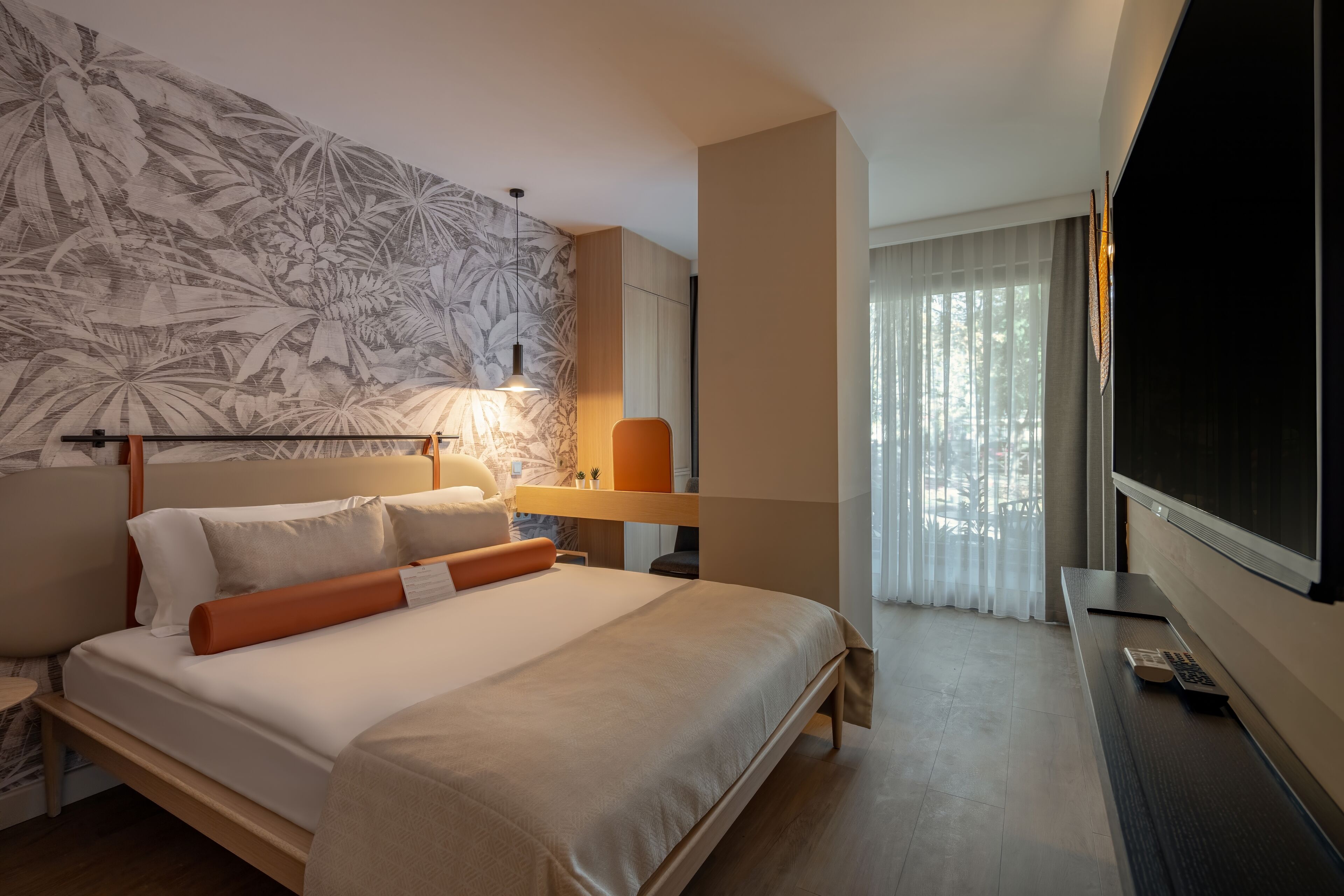 Una habitación de hotel moderna con una cama grande, un escritorio con lámpara, televisión y ventana con cortinas traslúcidas.