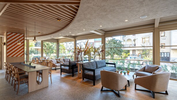 A spacious lobby with a wooden ceiling, large windows offering views of the street, a central seating area with sofas and chairs, and a long table. - Oli Hotel And Suites (Konyaalti)