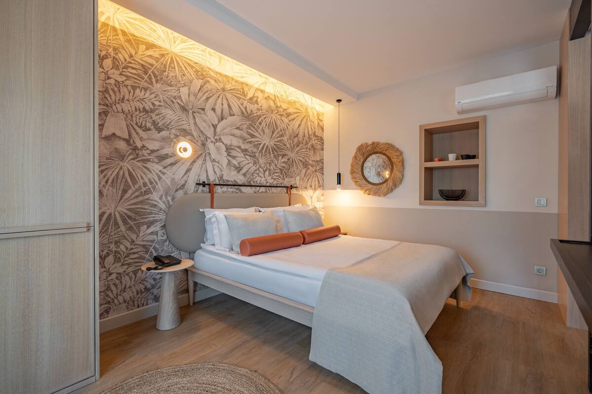 A bedroom with a bed, bedside table, wall-mounted shelf, and a decorative mirror.