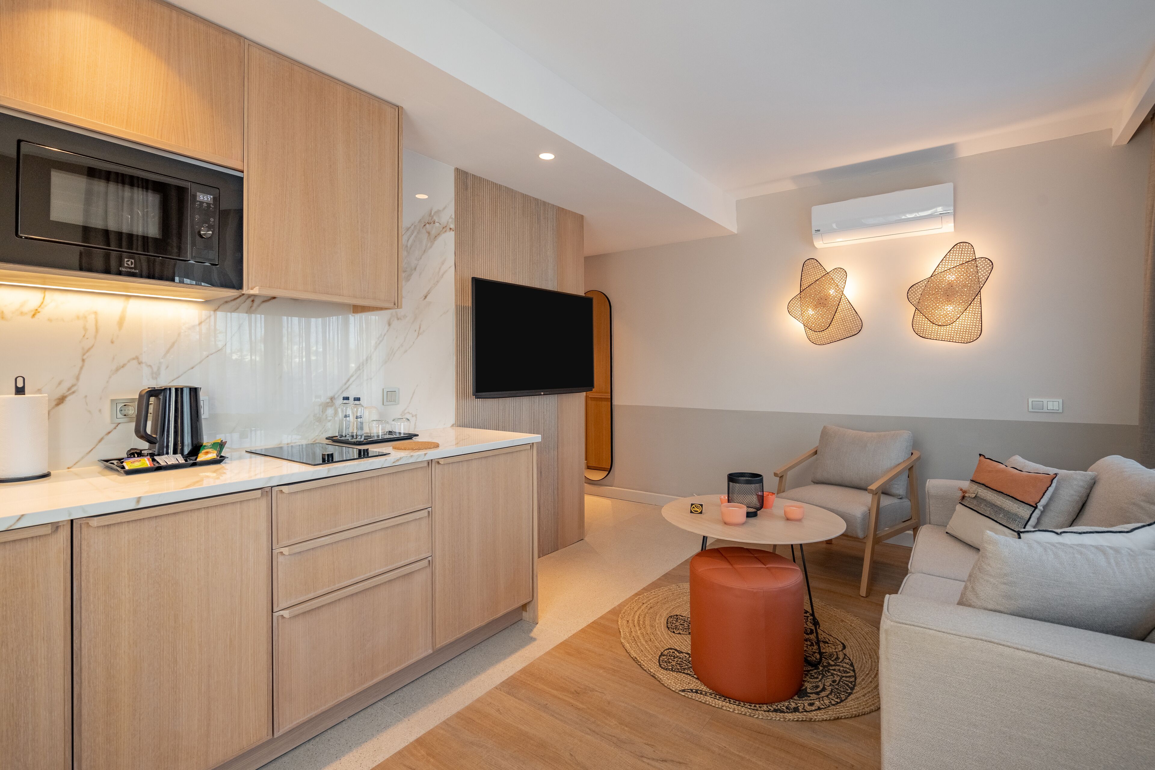 A modern kitchen with a microwave, stove, and sink. There is a flat-screen TV mounted on the wall, a small round table with chairs, and a sofa with cushions.