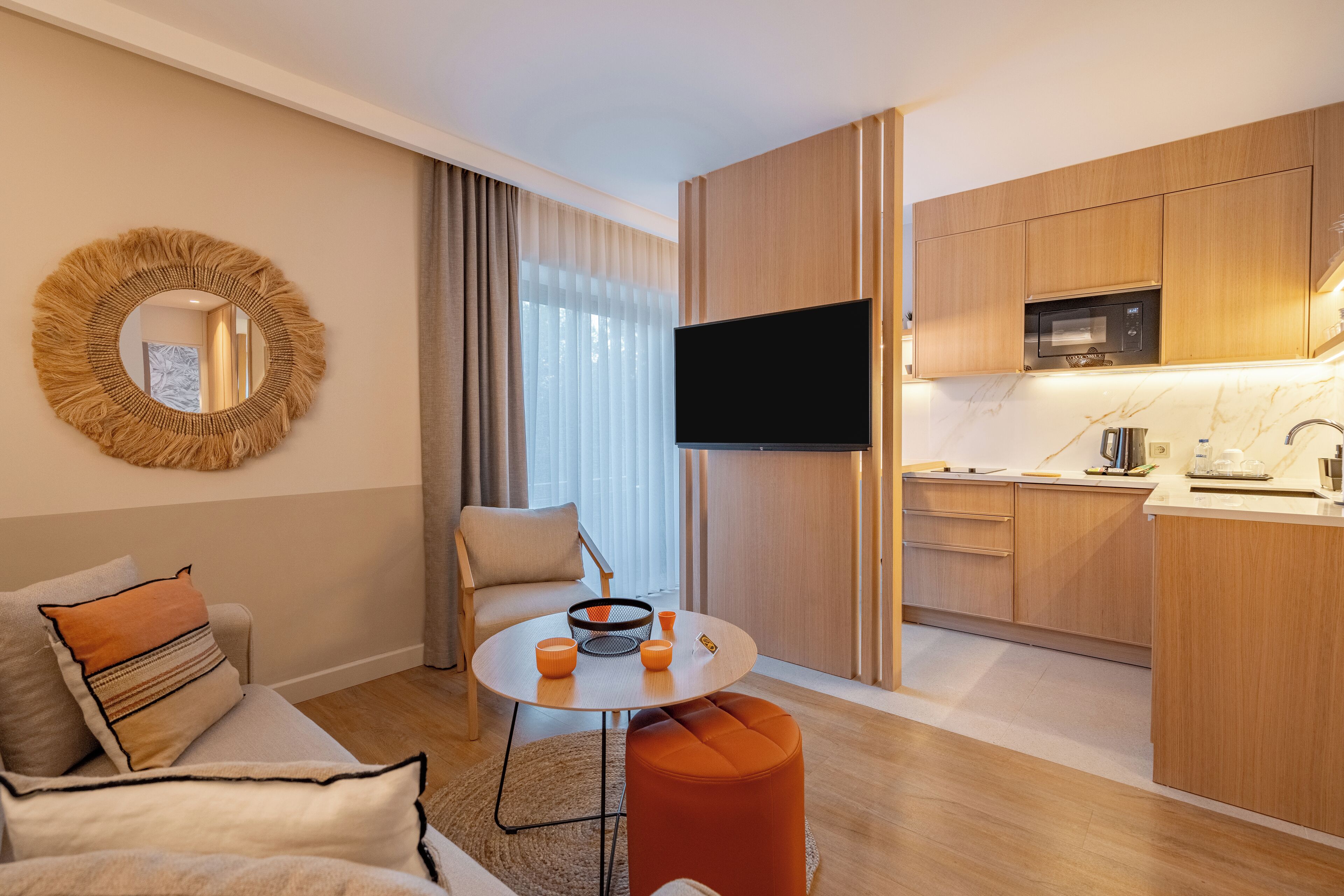 A modern hotel room with a round dining table, a sofa, a TV, and a kitchen area with wooden cabinets and a microwave.