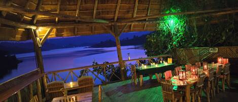 An outdoor dining area with wooden furniture and tables, overlooking a body of water at night.