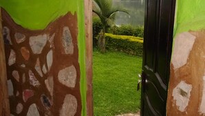Una habitación con pared de piedra, puerta verde y vista a un jardín bien cuidado con una palmera.