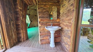 Una cabaña rústica de madera con piso de madera, un lavabo blanco y techo de paja visible a través de un umbral abierto.