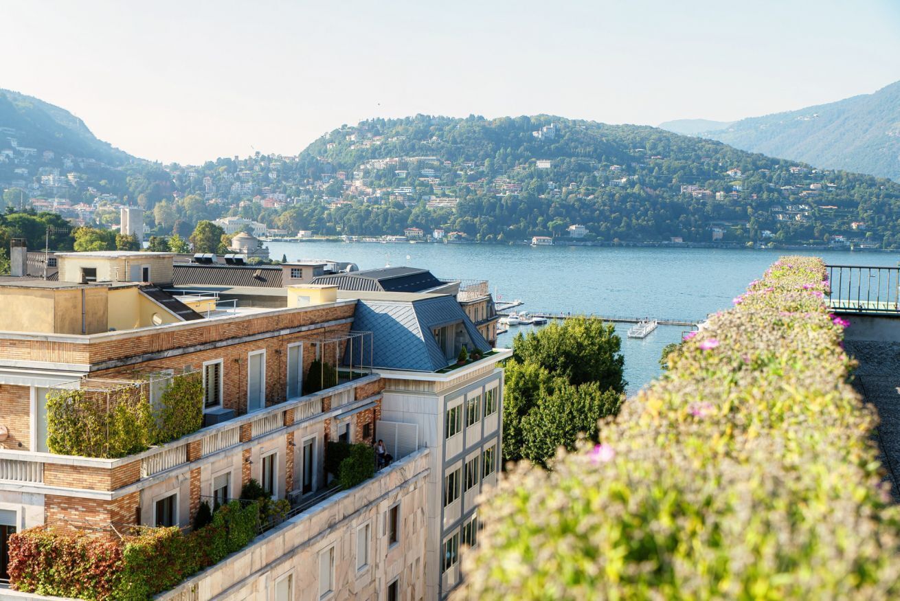 A multi-story building with a rooftop terrace, a view of a lake, and a hillside with buildings in the background.