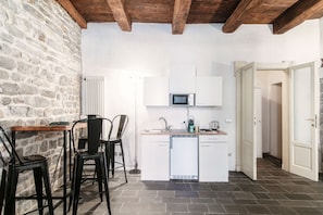 A compact kitchen with white cabinets, a microwave, and a dishwasher. There is a wooden table with chairs and a stone wall. - Angel's - My Home In Como (Como)