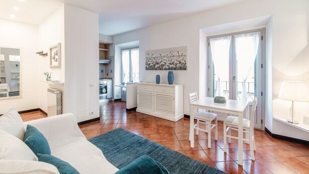 A modern living room with a sofa, a dining area with a table and chairs, a kitchen in the background, and a large window with curtains. - Celeste - My Home In Como (Como)