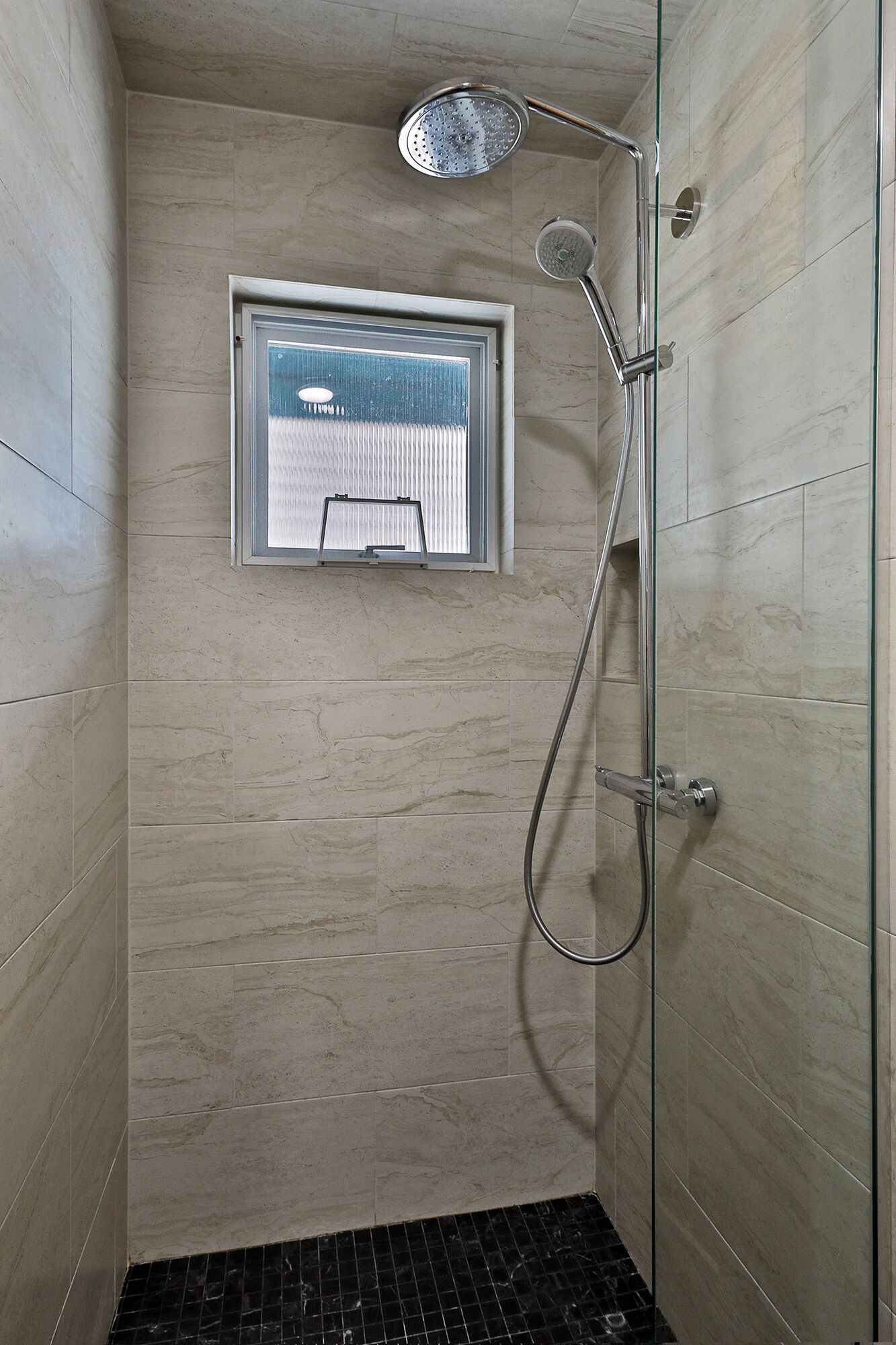 Une douche avec une porte en verre, une fenêtre et un sol carrelé.