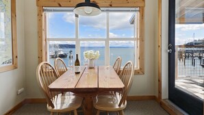 A dining area with a wooden table and chairs, a vase with flowers, and a view of the water through large windows.