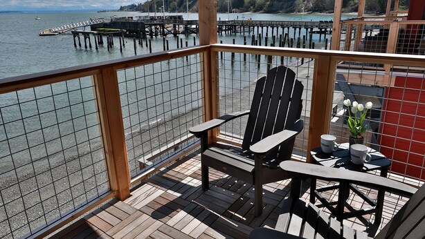 A wooden deck with a view of a dock and water. There are two Adirondack chairs and a small table with cups and a plant.