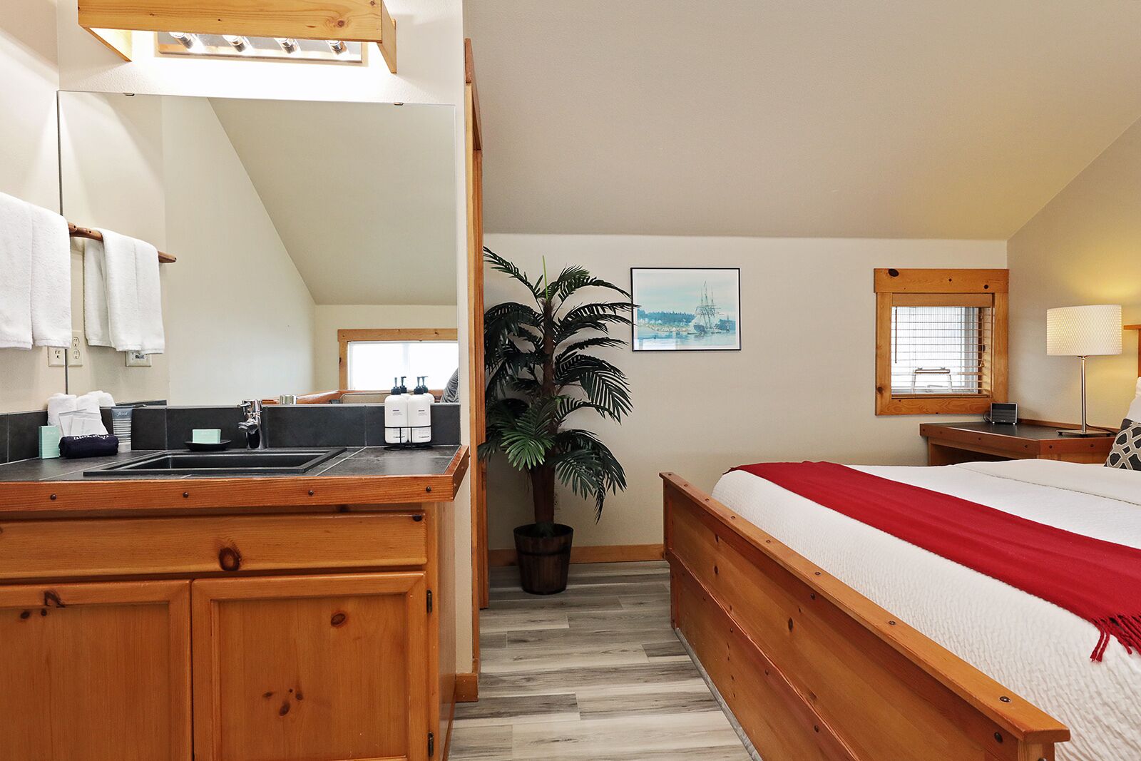 Habitación de hotel con una cama de madera, una planta grande en maceta, una zona de cocina con lavabo y armarios, y una ventana con persianas.