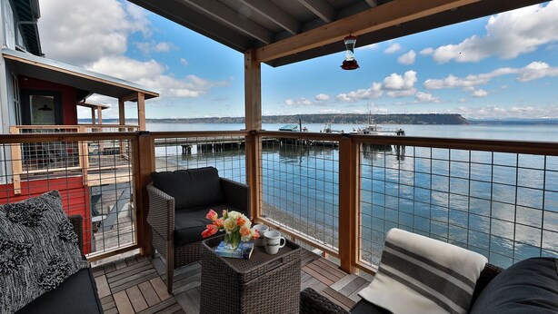 A covered outdoor deck with wicker furniture, a view of the water, and a dock in the background.