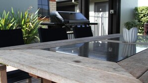 Mesa de vidrio con banco de madera, rodeada de sillas, en un entorno exterior con una parrilla y plantas en macetas.