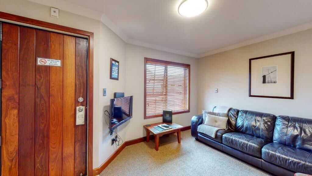 A living room with a wooden door, a black leather couch, a small table with a laptop, and a window with blinds.