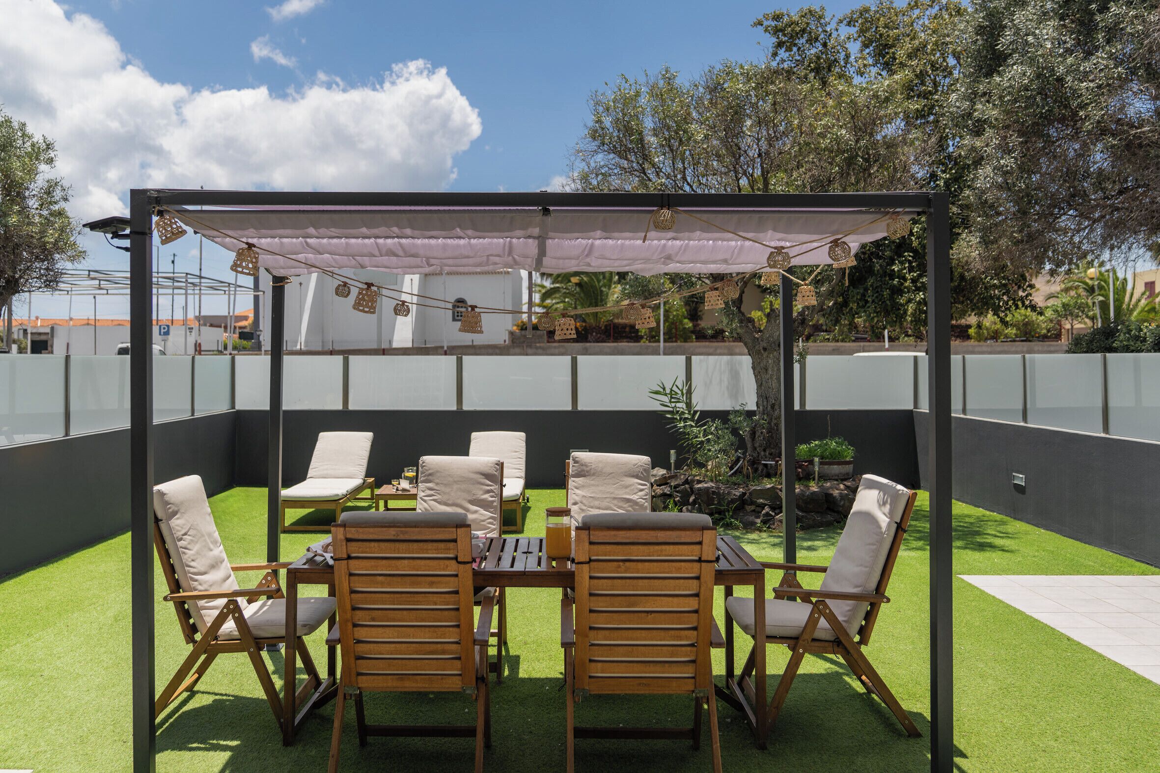 Een terras buiten met een pergola, zes houten stoelen en een tafel.
