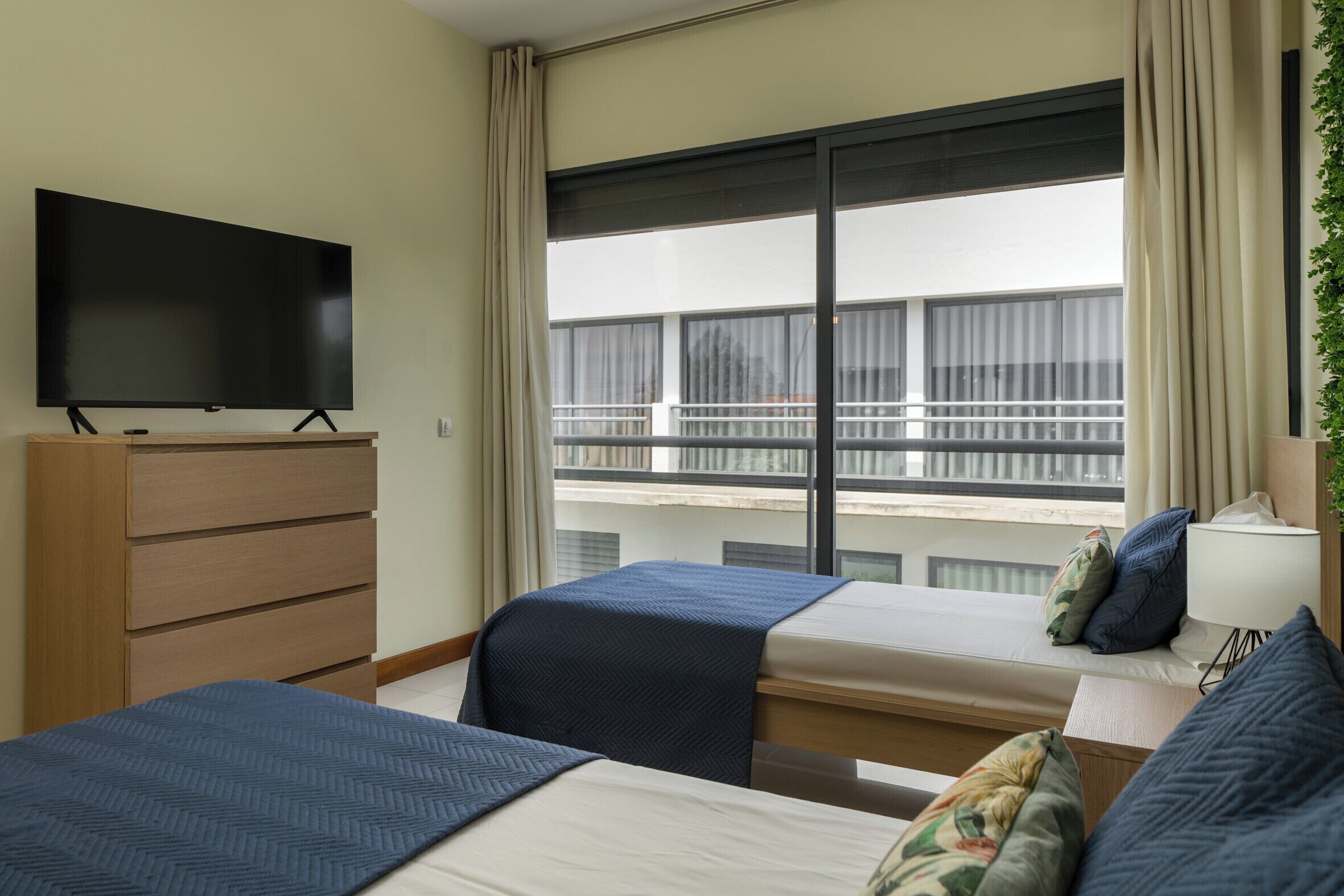 Ein Hotelzimmer mit Fernseher auf einer Holzkommode, zwei Betten mit blauer Bettwäsche, einem großen Fenster mit Blick auf Gebäude und einer Pflanze auf der rechten Seite.