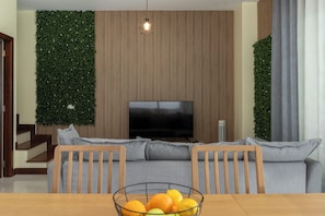 A modern living room with a flat-screen TV, a wooden dining table with a fruit bowl, and a sofa with grey cushions. - Vacation home in Porto Santo, Villa Girassol (Porto Santo)