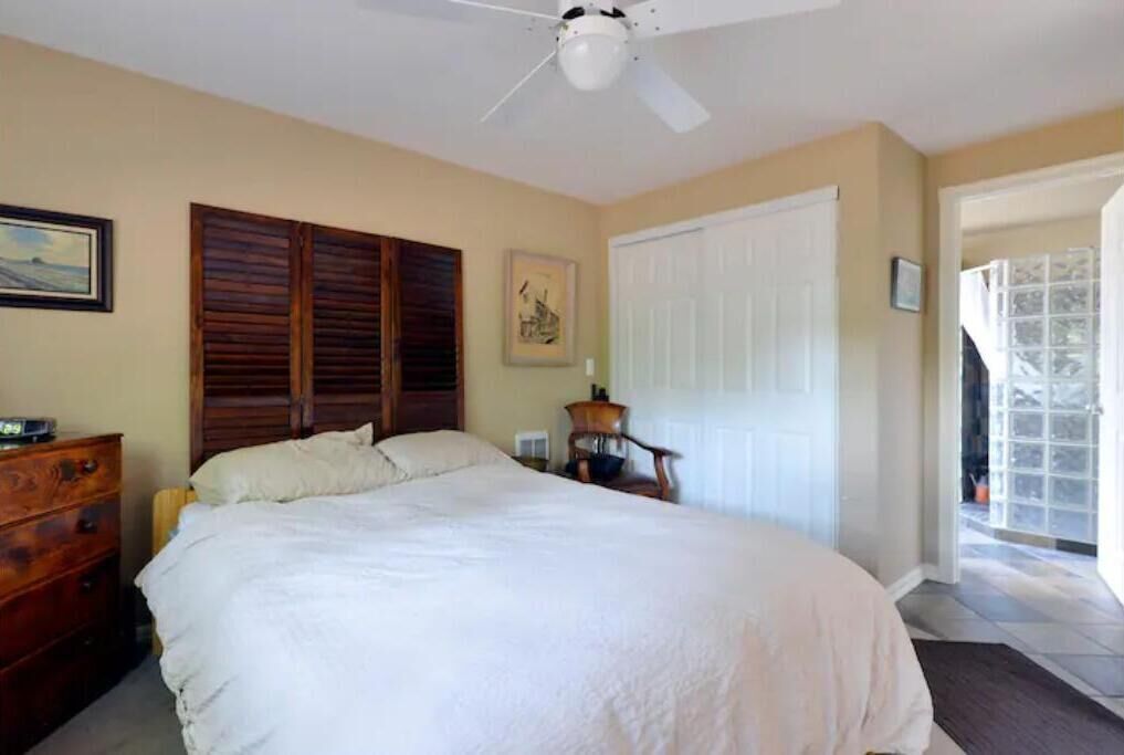 A bedroom with a bed, wooden headboard, a ceiling fan, a dresser, a chair, a door, and a window.