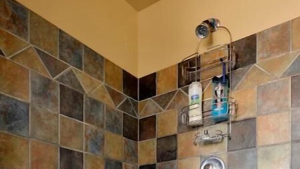 A shower area with a tiled wall and floor, a showerhead, and a metal shelf with toiletries. - Amazing Home...Large Space Yet Cozy (Kirkland)
