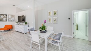Un salon moderne avec une table à manger blanche, des chaises blanches et un canapé orange. Des reproductions botaniques encadrées ornent le mur, et une plante en pot est posée sur la table.