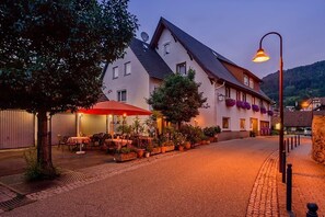 Ein Gebäude mit geneigtem Dach und mehreren Fenstern, umgeben von Blumentöpfen und Außenbestuhlung.
