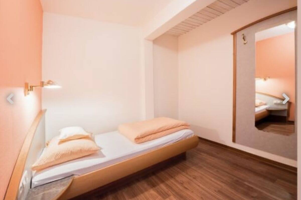 A small, neatly arranged bedroom with a single bed, a wooden floor, a mirror, and a wall-mounted light fixture.