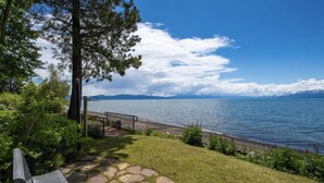 A lakeside view with a stone pathway leading to a bench, surrounded by greenery and a metal railing.