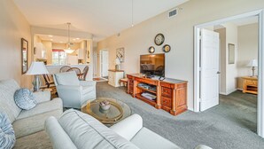 A spacious living room with a sofa, coffee table, and TV stand. There is a dining area in the background with a chandelier. - Naples Condo w/ Pond Views & Patio - Near Golf! (Naples)