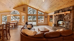 A cozy cabin interior with a stone fireplace, wooden ceiling, large windows, and a leather sofa.