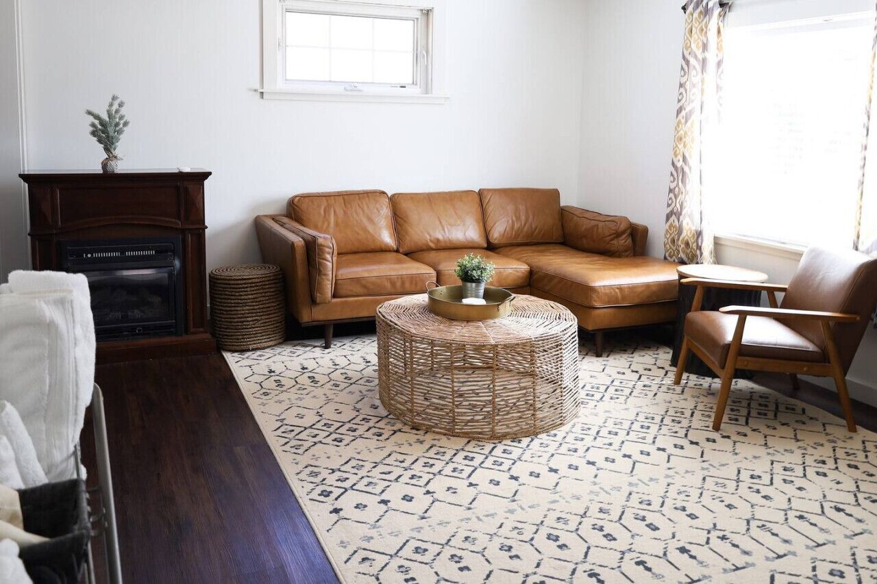 A living room with a brown leather sofa, a round wicker coffee table, a wooden side table, a fireplace, and a patterned rug.