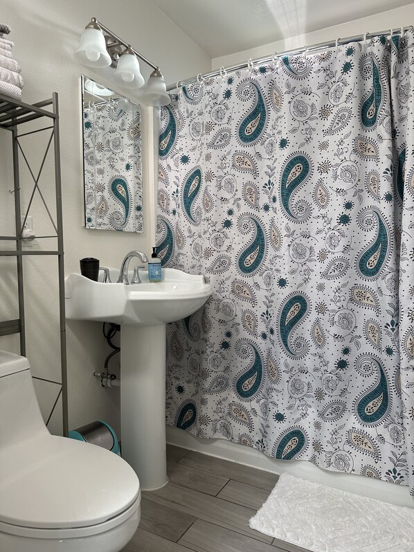 A bathroom with a white sink, a mirror above it, a shower curtain with paisley patterns, and a white towel on the floor.