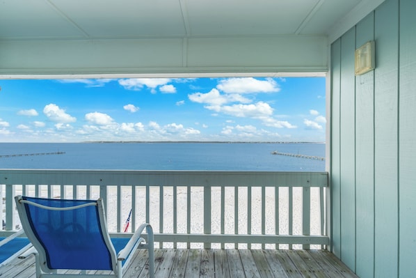 A wooden deck with a blue chair, a white railing, and a view of the beach and ocean.