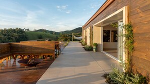 A wooden deck with outdoor furniture, a concrete pathway, and a building with large windows and greenery. - Vista Deslumbrante e Alto Padrão com 4 Suites, Piscina Aquecida e Sauna (Santo Antônio do Pinhal)
