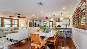 A spacious living area with a dining table, wicker chairs, and a sofa. A kitchen with blue cabinets and a bar area is visible in the background.