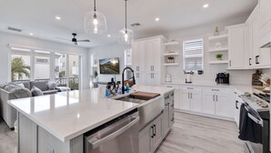 Una cocina moderna con armarios blancos, una isla grande con lavavajillas, luces colgantes y vistas al exterior.