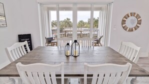 Un comedor con mesa de madera y sillas blancas, dos faroles sobre la mesa y vistas a palmeras afuera.