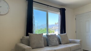 A living room with a sofa, a clock on the wall, and a view of mountains through the window. - CocoVillaSB Apartment #4 (Santa Barbara)