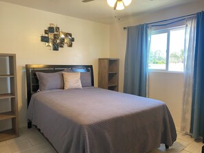Un dormitorio con cama, ventilador de techo, un adorno en la pared y una ventana con cortinas.